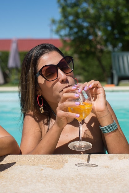 Pretty woman drinking bright cocktail in pool. Woman with dark hair holding glass with bright beverage, looking at camera. Leisure, friendship, party concept