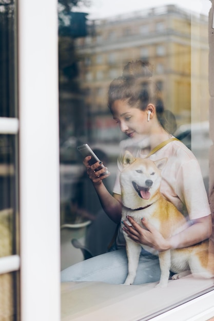 Pretty woman in earphones using smartphone hugging dog in cafe