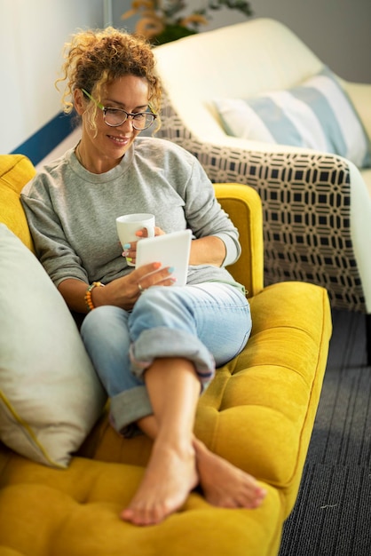 La donna graziosa si diverte a rilassarsi a casa leggendo un libro sul lettore da tavolo e bevendo un tè o un caffè