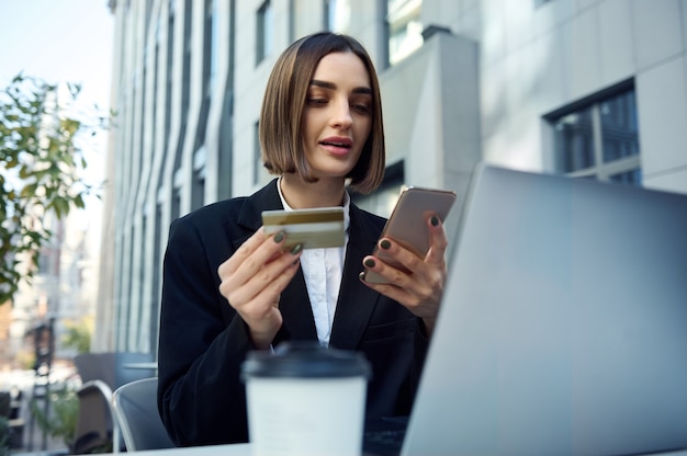 Pretty woman, entrepreneur, copywriter holding mobile phone and gold credit card sitting at table in outside public park and working on laptop, shopping online, booking. Remote business concept