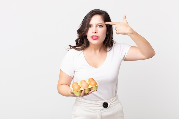 Pretty woman feeling confused and puzzled, showing you are insane and holding an eggs box