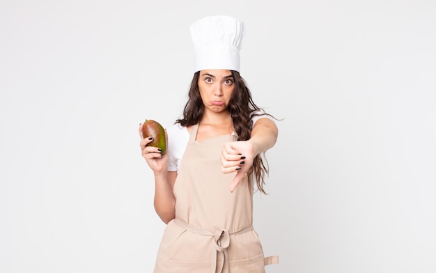 Pretty woman feeling crossshowing thumbs down wearing an apron and holding a mango