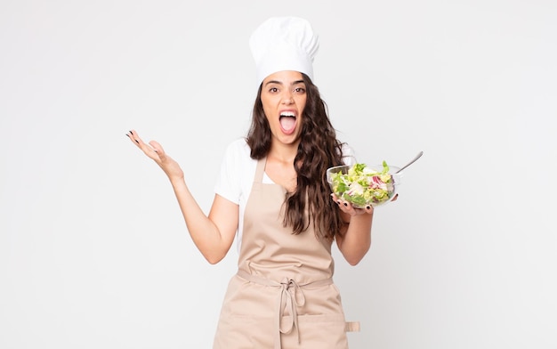 Pretty woman feeling extremely shocked and surprised wearing an apron and holding a salad