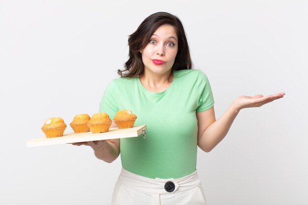 Pretty woman feeling puzzled and confused and doubting and holding a muffins tray