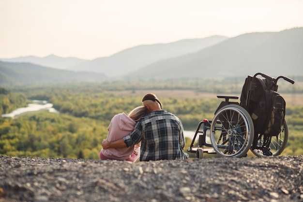 Pretty woman and her incapacitated husband resting together near his wheelchair on the hill.