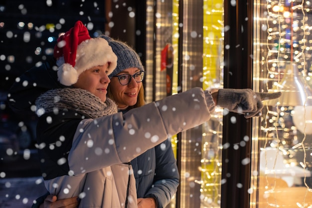 Photo pretty woman and her son in santas hat looking and dreaming in illuminated shop window