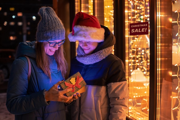 Pretty woman and her son in santas hat opening gift box on illuminated shop window background. New Year or Christmas concept