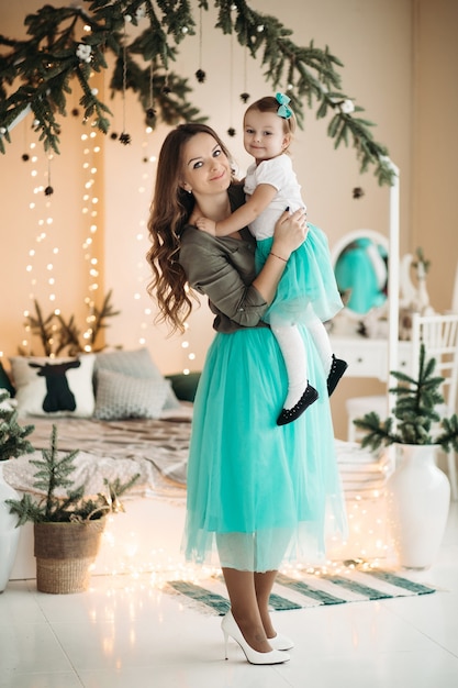 Pretty woman holding daughter in hands, standing in decorated studio with fir branches and light garland