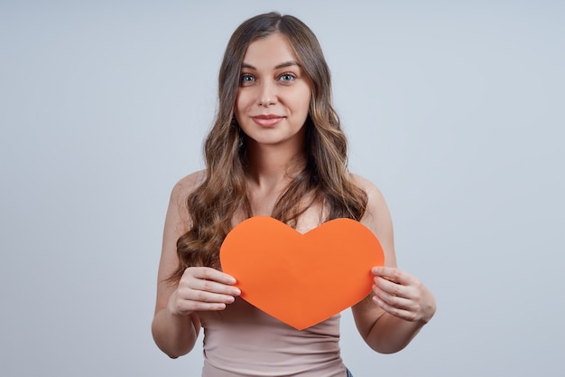 Photo pretty woman holding a paper heart at her chest, looking at the camera, on gray background. happy valentine's day. world heart day.