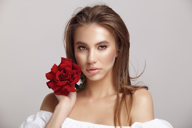 Pretty woman holds red rose flower by chin looking at camera