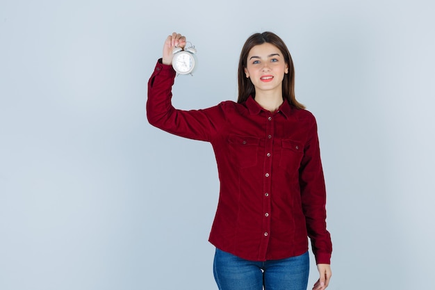 Pretty woman keeping alarm clock while smiling in burgundy blouse and looking joyous.