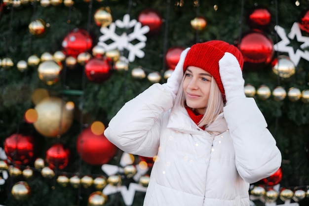 ニットの赤い帽子と白いコートを着たきれいな女性が街のクリスマスツリーに立っています