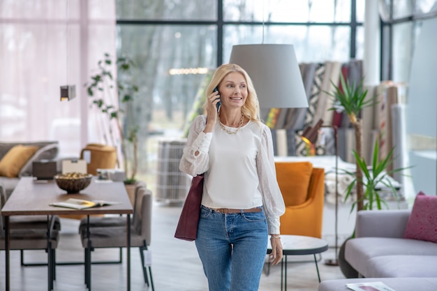 Pretty woman in a light blouse talking on a smartphone, looking to the side walking through a furniture store, smiling.