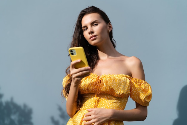 Pretty woman long dark hair in bright yellow summer dress posing on blue wall background daylight holding mobile phone and looking to camera