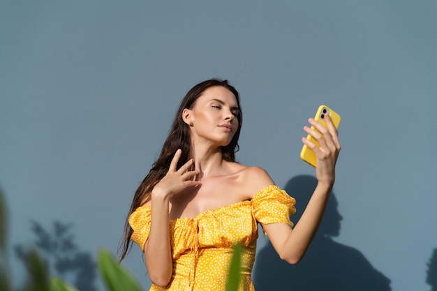 Pretty woman long dark hair in bright yellow summer dress posing on blue wall background daylight taking selfie on mobile phone