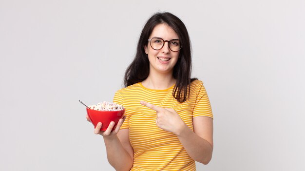 Pretty woman looking excited and surprised pointing to the side and holding a breakfast bowl