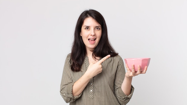 Pretty woman looking excited and surprised pointing to the side holding an empty pot bowl. assistant agent with a headset