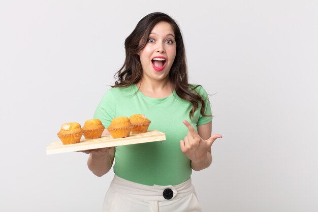 Pretty woman looking excited and surprised pointing to the side and holding a muffins tray