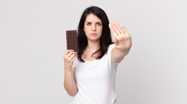 Pretty woman looking serious showing open palm making stop gesture and holding a chocolate bar