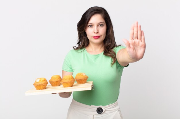 Pretty woman looking serious showing open palm making stop gesture and holding a muffins tray