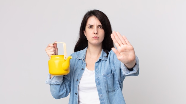 Pretty woman looking serious showing open palm making stop\
gesture and holding a teapot