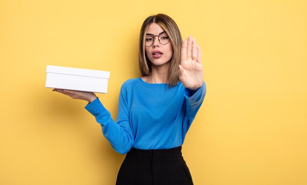 Pretty woman looking serious showing open palm making stop gesture. holding white box