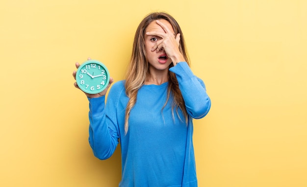 Photo pretty woman looking shocked scared or terrified covering face with hand holding an alarm clock