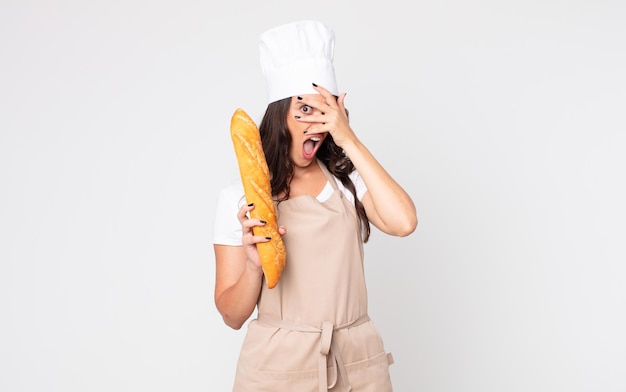 Pretty woman looking shocked, scared or terrified, covering face with hand wearing an apron and holding a bread baguette