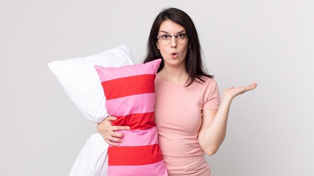 Pretty woman looking surprised and shocked, with jaw dropped holding an object wearing pajamas and holding a pillow