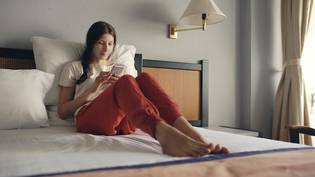 Pretty woman lying in bed in hotel room and using smartphone and smiling