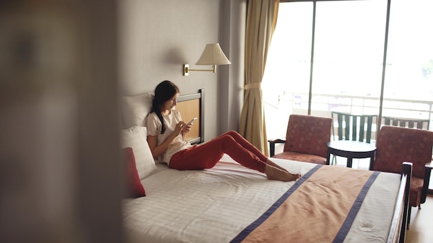 Foto bella donna sdraiata a letto nella camera d'albergo e utilizzando smartphone e sorridente