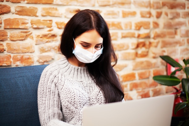 Pretty woman in mask on her face working on laptop at home during pandemic