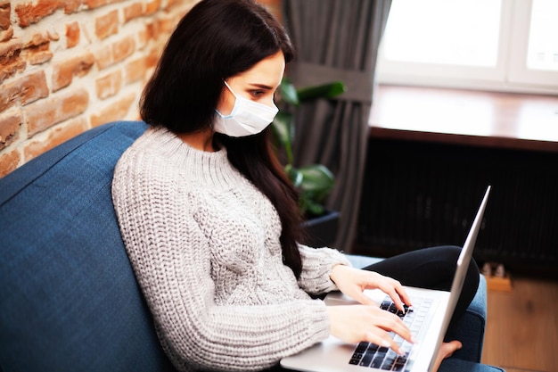 Pretty woman in mask on her face working on laptop at home during pandemic.