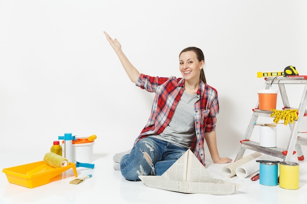 Pretty woman pointing hand aside on copy space, sit on floor with newspaper hat, instruments for renovation apartment isolated on white background. Wallpaper, painting tools. Repair home concept.