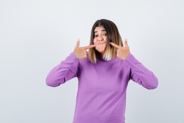 Pretty woman pointing at her puffy cheeks in purple sweater and looking playful , front view.