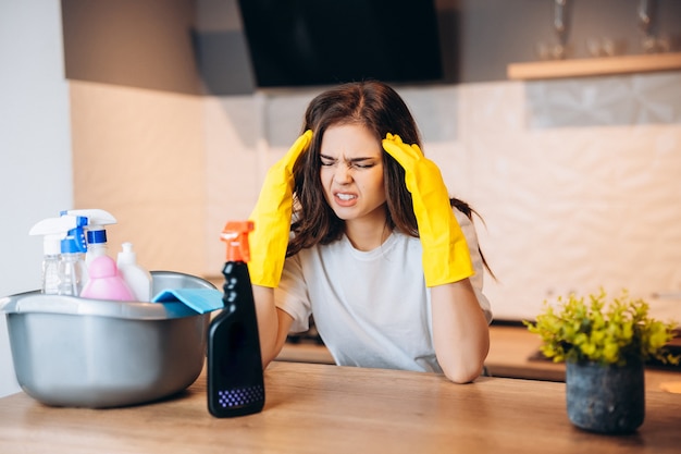 Pretty woman in protective yellow gloves is having headache in the kitchen while cleaning and totally doesn't like it