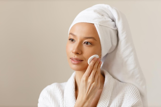 Pretty woman in robe and towel on her head wipes the skin of her face with cotton pads Cleansing and healthy body care after shower Closeup of girl on isolated background