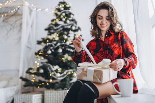 Pretty woman in shirt and socks sitting on the windowsill with Christmas gift box