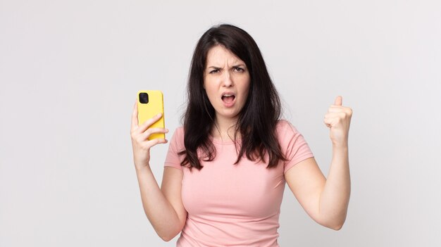 Красивая женщина агрессивно кричит с сердитым выражением лица, используя смартфон