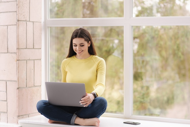 Pretty woman sitting on window sill with laptop and woking. Freelancer at home barefoot. Brunette female in yellow sweatshirt.