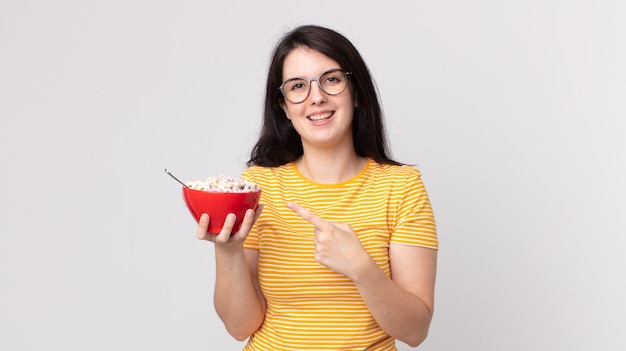 Pretty woman smiling cheerfully, feeling happy and pointing to the side and holding a breakfast bowl