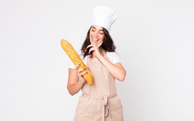 pretty woman smiling happily and daydreaming or doubting wearing an apron and holding a bread baguette