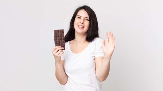 Красивая женщина счастливо улыбается, машет рукой, приветствует и приветствует вас и держит плитку шоколада