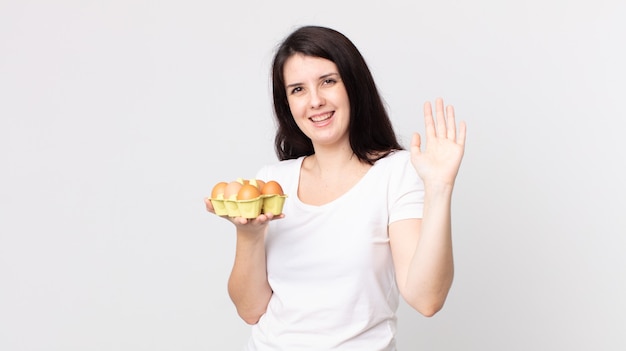 Pretty woman smiling happily, waving hand, welcoming and greeting you and holding an eggs box