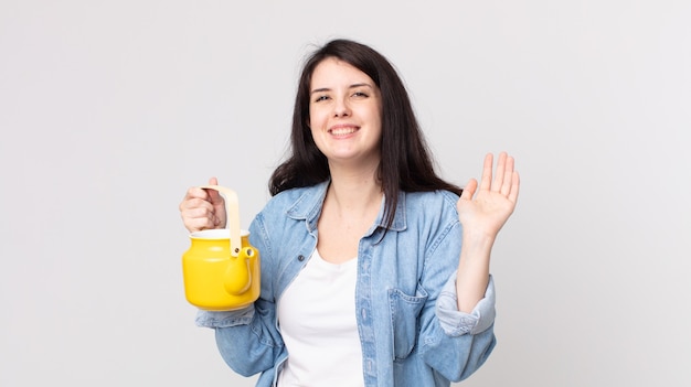 Pretty woman smiling happily, waving hand, welcoming and greeting you and holding a teapot