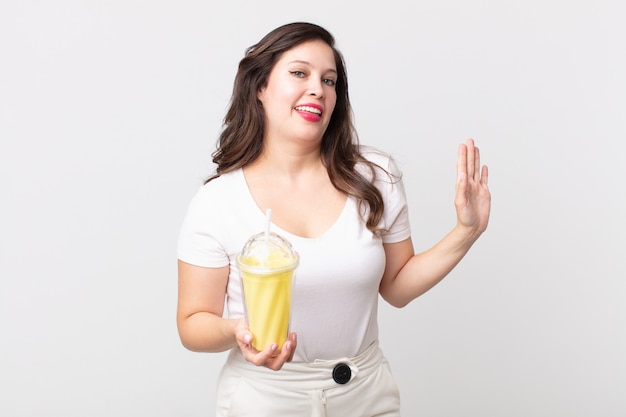 Красивая женщина счастливо улыбается, машет рукой, приветствует и приветствует вас и держит ванильный молочный коктейль