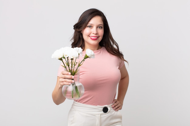 Pretty woman smiling happily with a hand on hip and confident and holding a decorative flowers pot