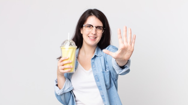 Pretty woman smiling and looking friendly, showing number five and holding a vanilla milkshake