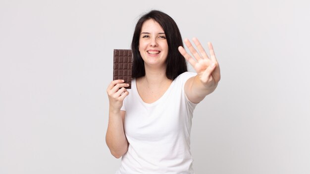 Pretty woman smiling and looking friendly, showing number four and holding a chocolate bar