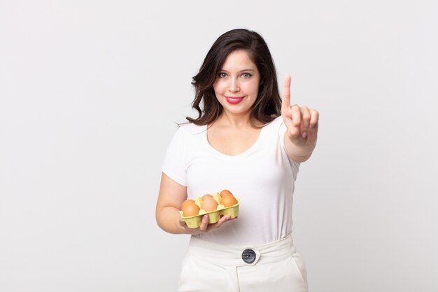 Pretty woman smiling and looking friendly, showing number one and holding an eggs box